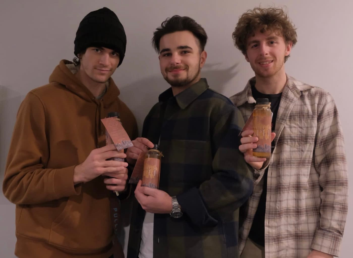 Three young men standing against a white wall, each holding a bottle of Ikigai kombucha with an orange label.